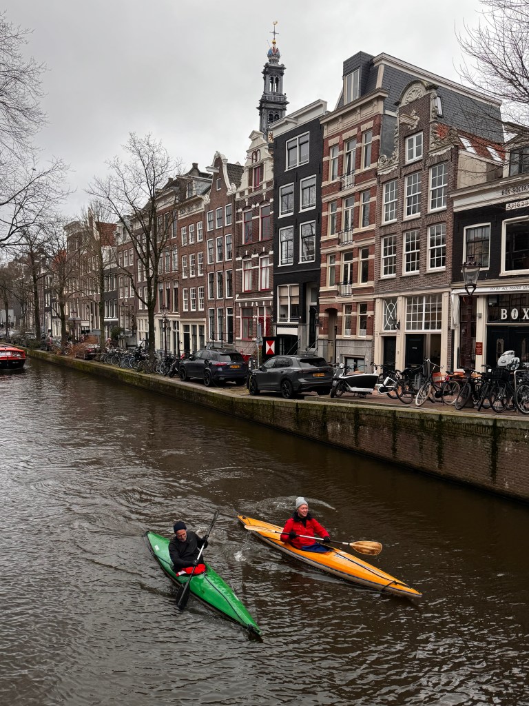 Two kayakers paddling in a canal surrounded by historic buildings in Amsterdam, with a cloudy sky overhead.