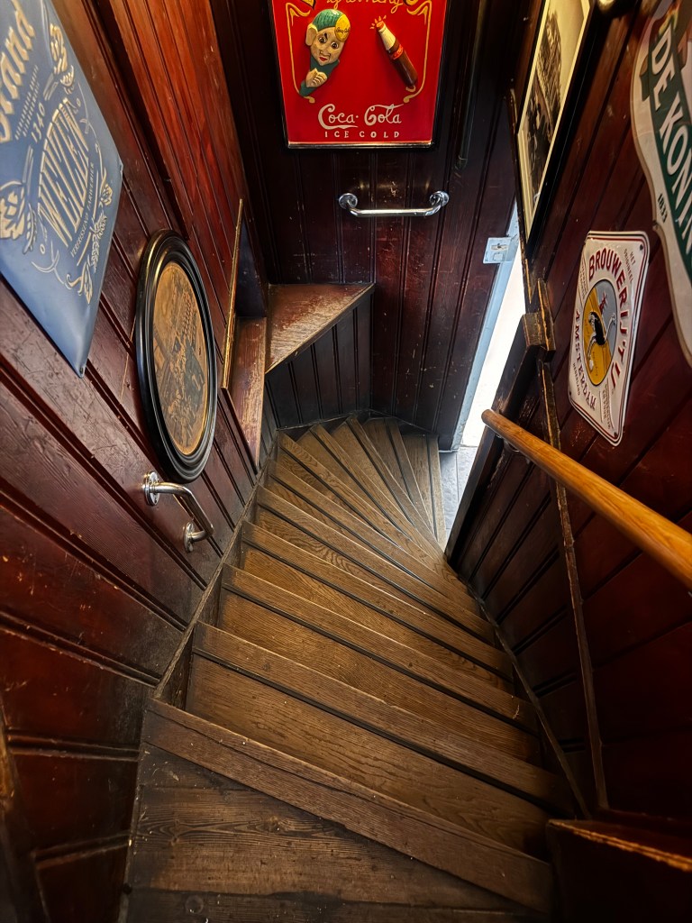 A downward view of a spiraled wooden staircase leading to an exit, surrounded by vintage signs and dark wooden walls.
