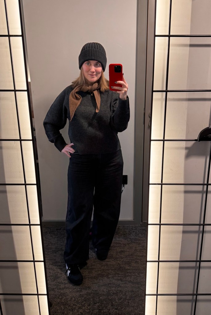 A woman stands in front of a mirror, wearing a black knit beanie and a grey sweater with a brown scarf tied around her neck. She is also dressed in loose-fitting black pants and black shoes, holding a red smartphone to take a selfie.