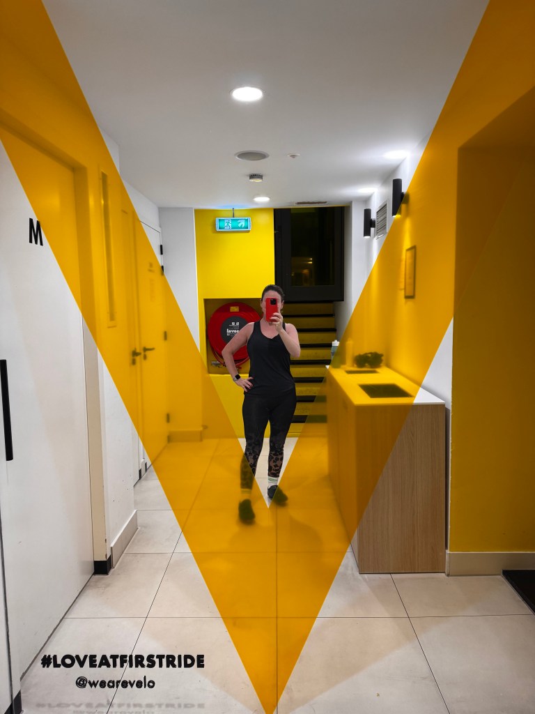 A person taking a selfie in a brightly colored hallway with yellow walls and a modern design, featuring a distinct geometric yellow shape and a staircase in the background. The person is wearing athletic clothing and holding a red phone.