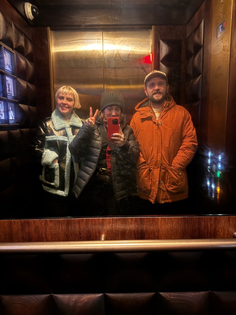 A group of three people posing together in an elevator, with one person in the center taking a selfie. The two individuals on either side are smiling and making peace signs, while the elevator features a stylish wooden interior and digital control panel.