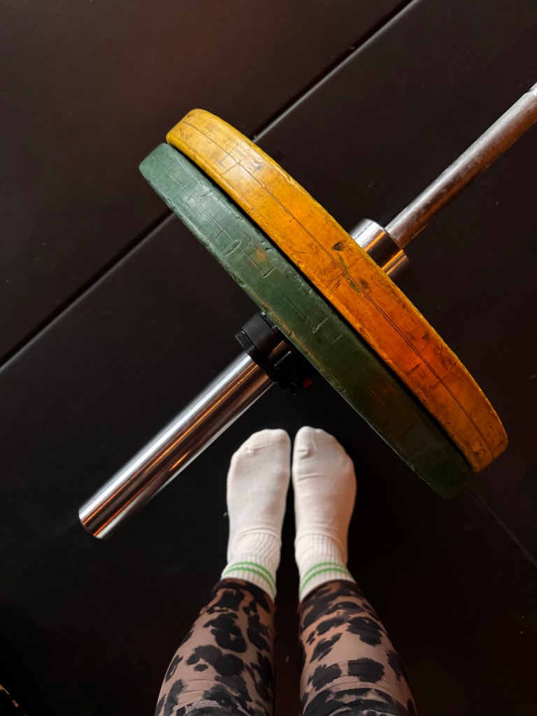 Feet wearing white socks standing next to a barbell with green and yellow weight plates on a gym floor.