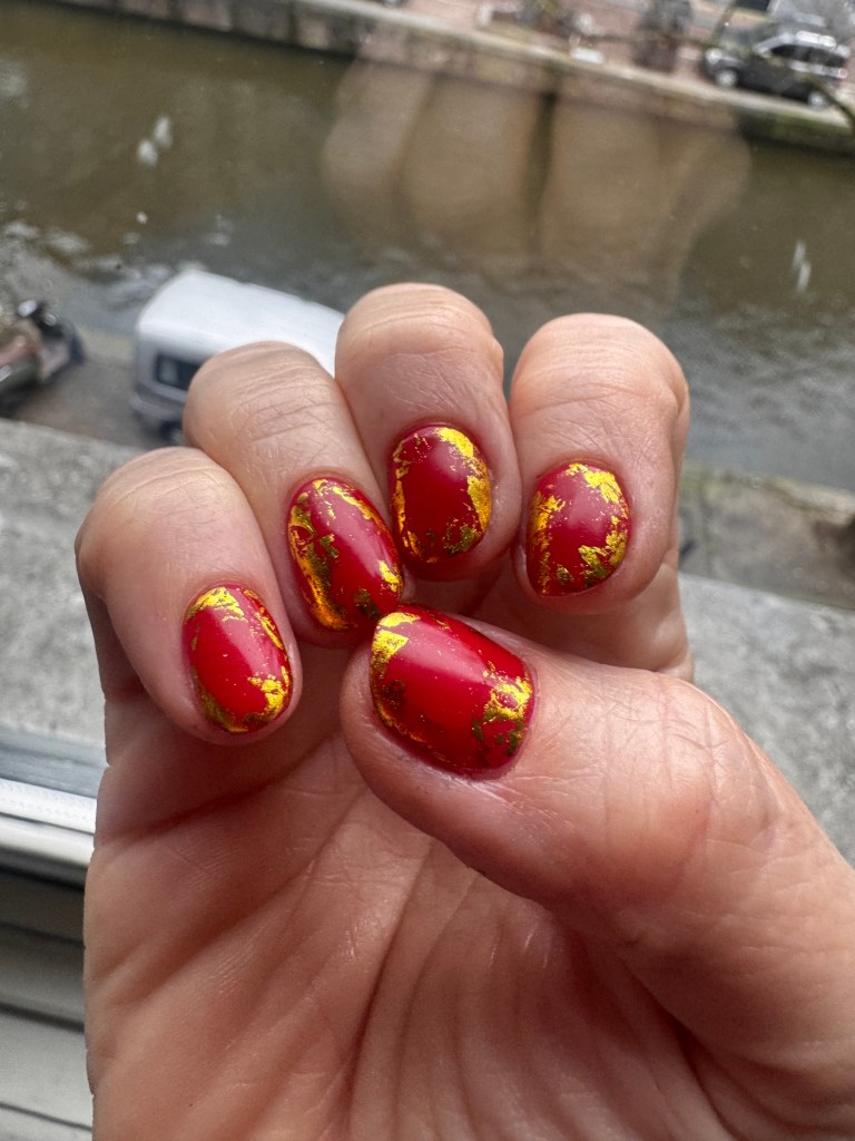 Close-up of a hand with beautifully manicured red nails adorned with gold leaf accents, set against a backdrop of a canal.