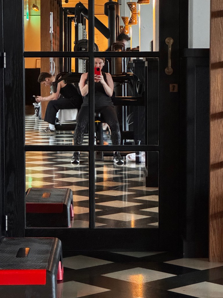 A gym scene reflecting through a glass partition, showing a person sitting on a bench taking a selfie with a phone, while another person is working out in the background.