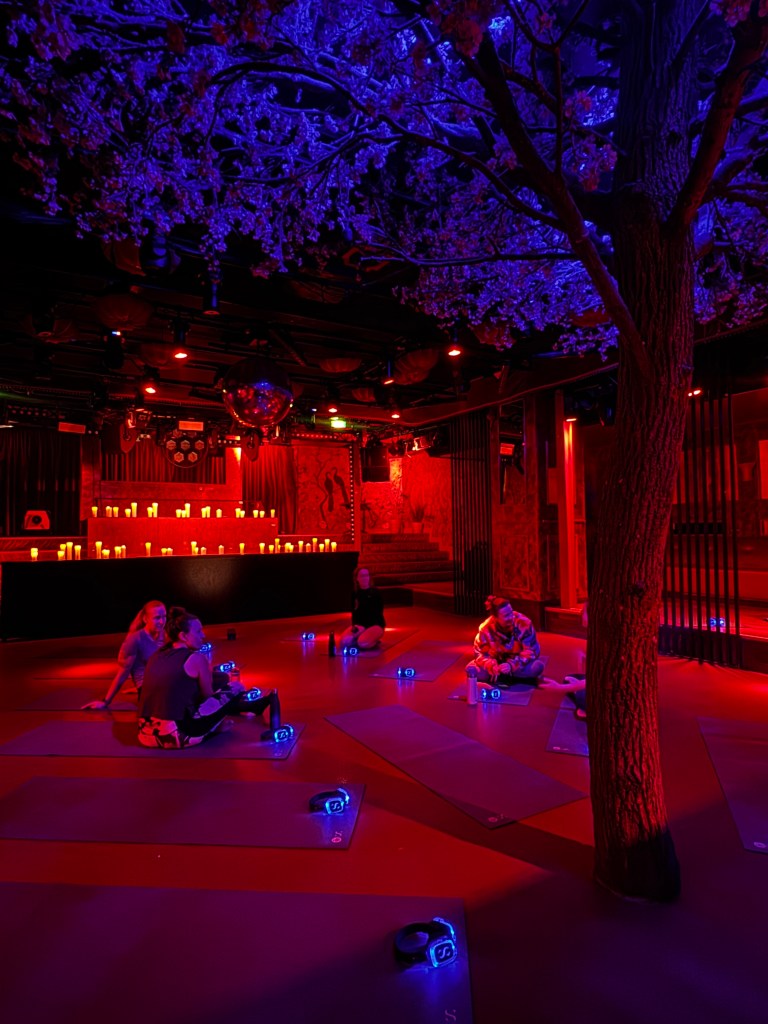 A dimly lit room with a purple and red ambiance featuring participants on yoga mats, surrounded by decorative cherry blossom branches and glowing candles.