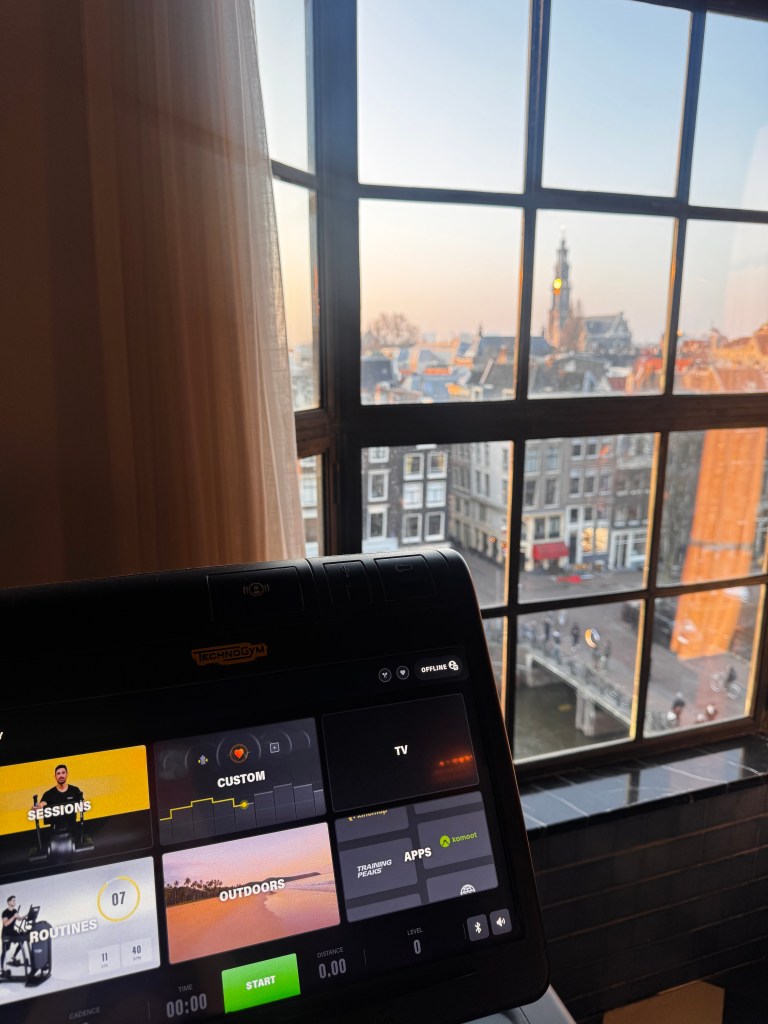 View from a gym window overlooking a cityscape at sunrise, with a treadmill interface displaying various workout options.