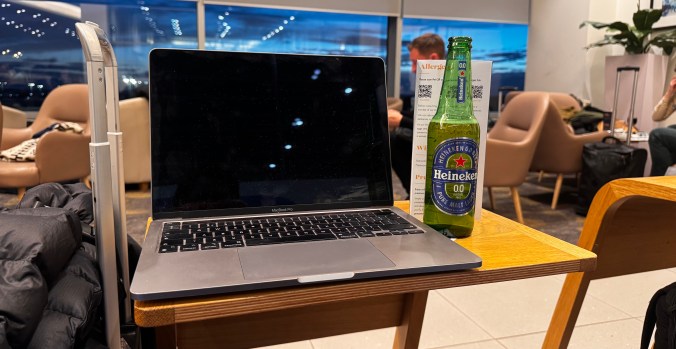 A laptop on a wooden table next to a Heineken 0.0 beer bottle, with a view of a lounge area in the background.