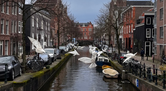 A scenic view of a canal in a city, lined with brick buildings and parked bicycles. Several boats are visible in the water, along with whimsical, white paper-like figures resembling birds, creating an artistic touch to the urban landscape.