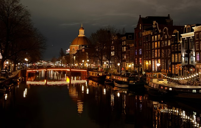 Night view of a canal in Amsterdam with illuminated buildings, boats, and a dome-shaped structure in the background.