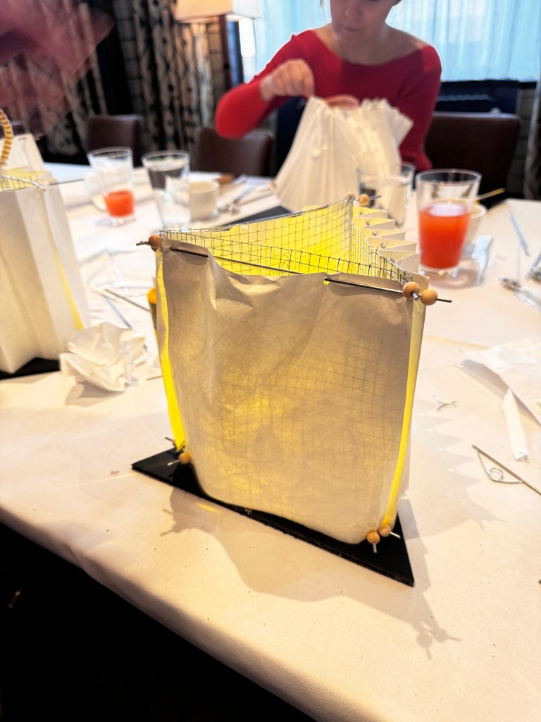 A partially assembled decorative lantern made of white paper with yellow accents, placed on a table set with glasses and utensils.