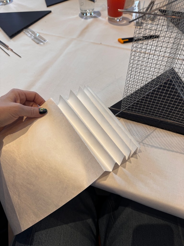 A hand holding a folded sheet of paper with multiple creases, set on a table with a wire sculpture, a glass of beverage, and various tools in the background.