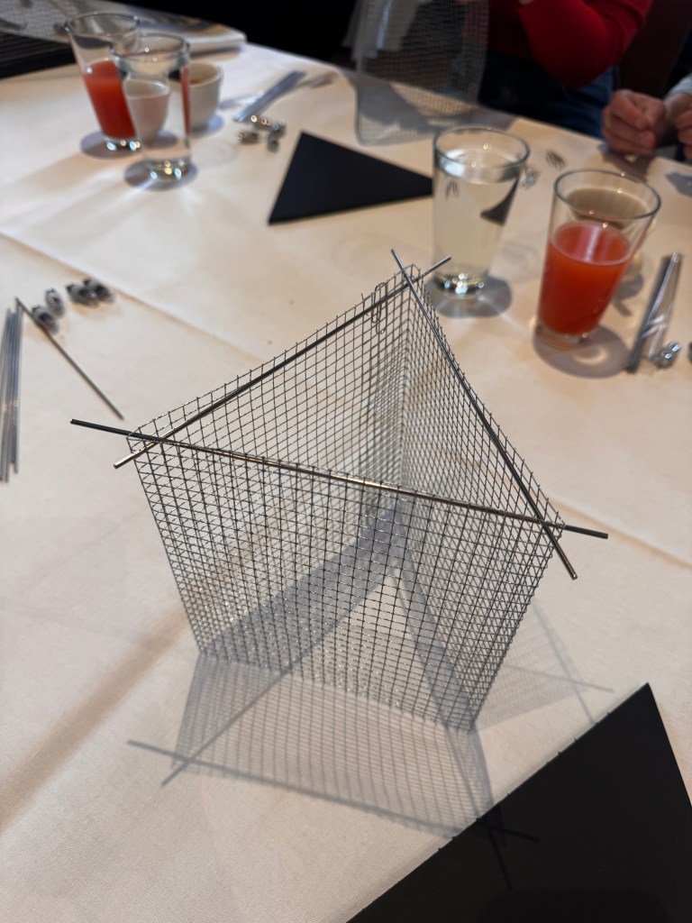 A metallic mesh sculpture resembling a pyramid, set on a table with various glasses and utensils in the background.