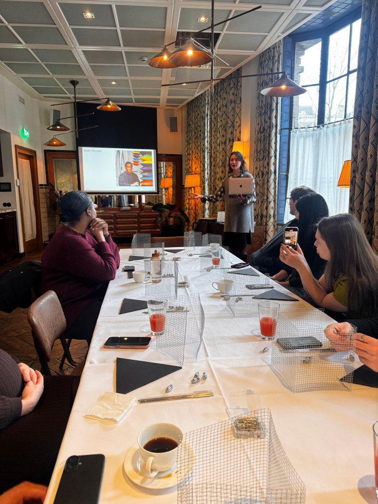 A woman presenting with a laptop in a meeting room, with a projector screen displaying an image, while attendees sit at a long table with drinks and notebooks.