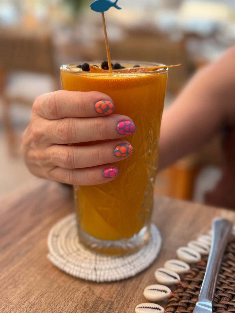 A hand holding a tall glass of orange drink adorned with a small fish decoration, featuring colorful nail art with geometric designs.