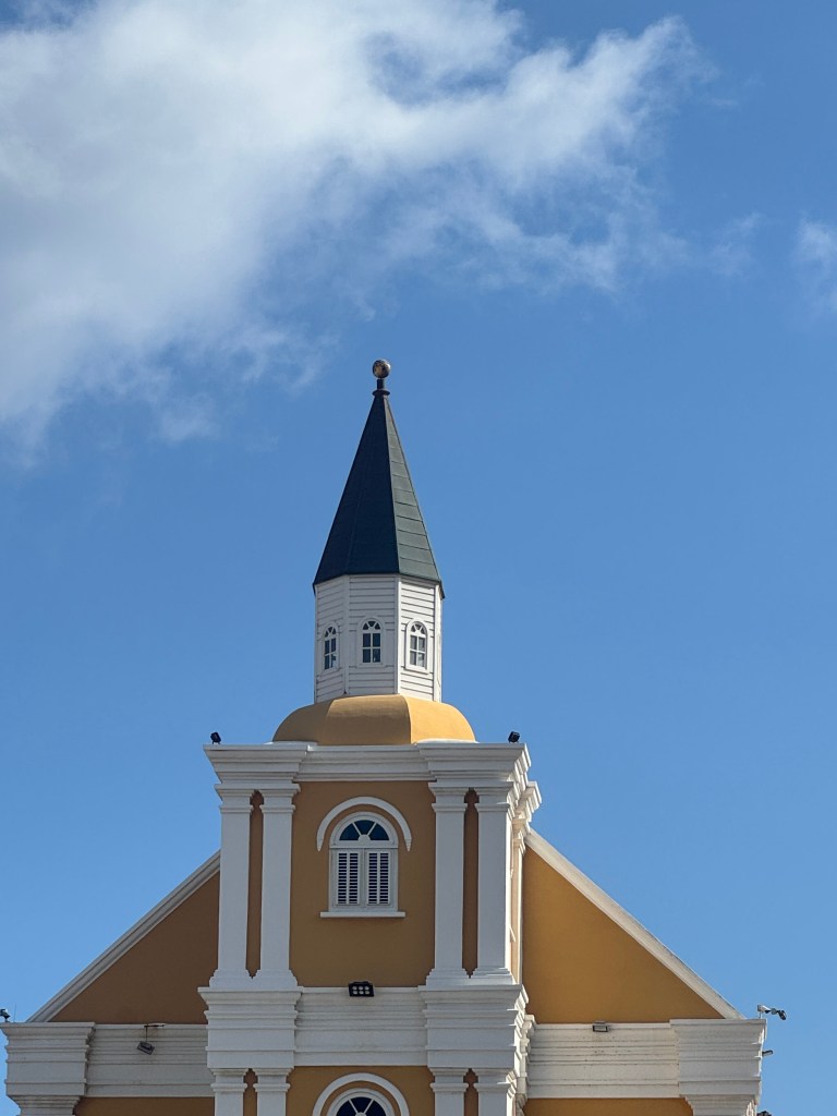 Close-up of the steeple of a yellow building with a pointed roof and decorative windows against a blue sky.