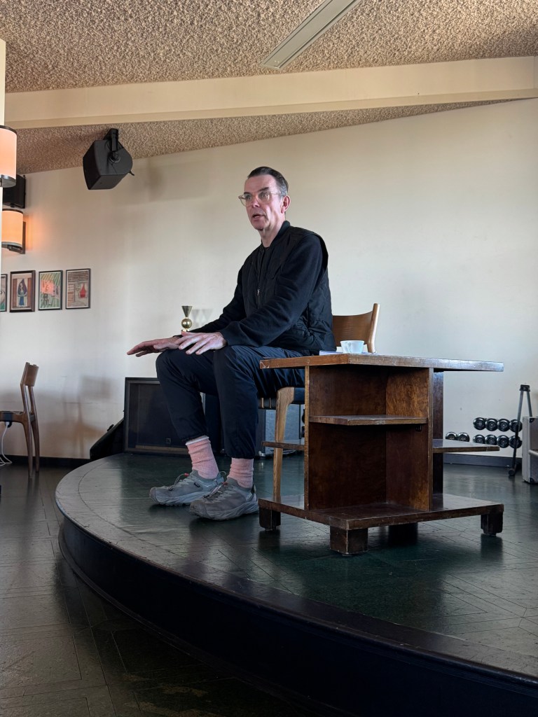 A person sitting on a wooden stool at a small table in a modern interior space, with dim lighting and decorative wall art.