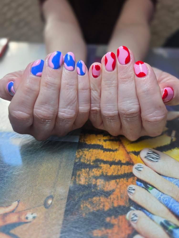 Close-up of two hands showing colorful nail art with blue, pink, and red swirls on the nails against a patterned background.
