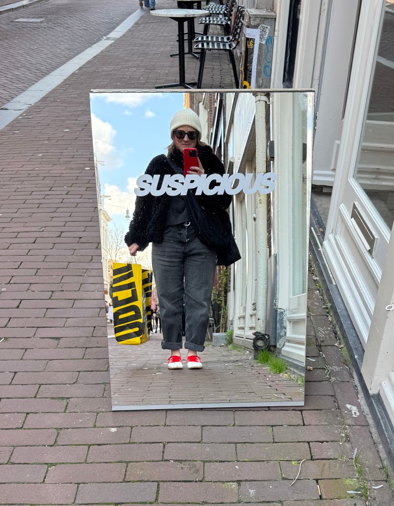 A person wearing a beanie and sunglasses is reflected in a mirror displaying the word 'SUSPICIOUS'. They are holding a bright yellow shopping bag and wearing red shoes while standing on a brick sidewalk.
