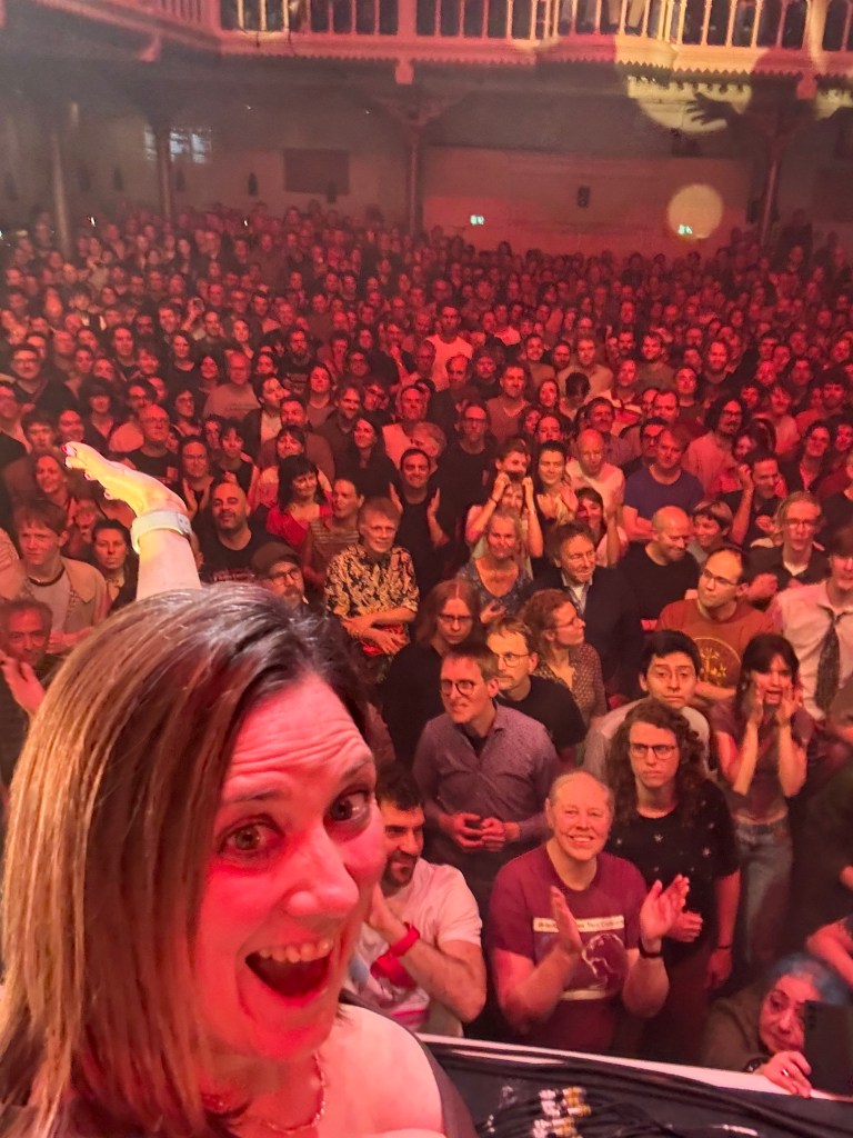 A lively concert scene featuring a large crowd of enthusiastic audience members, with a woman in the foreground taking a selfie, smiling excitedly.