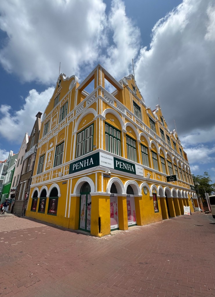 A large yellow building with arched windows and decorative accents, featuring a sign that reads 'PENHA'. The building has a classic architectural style and is situated on a brick-paved street under a partly cloudy sky.