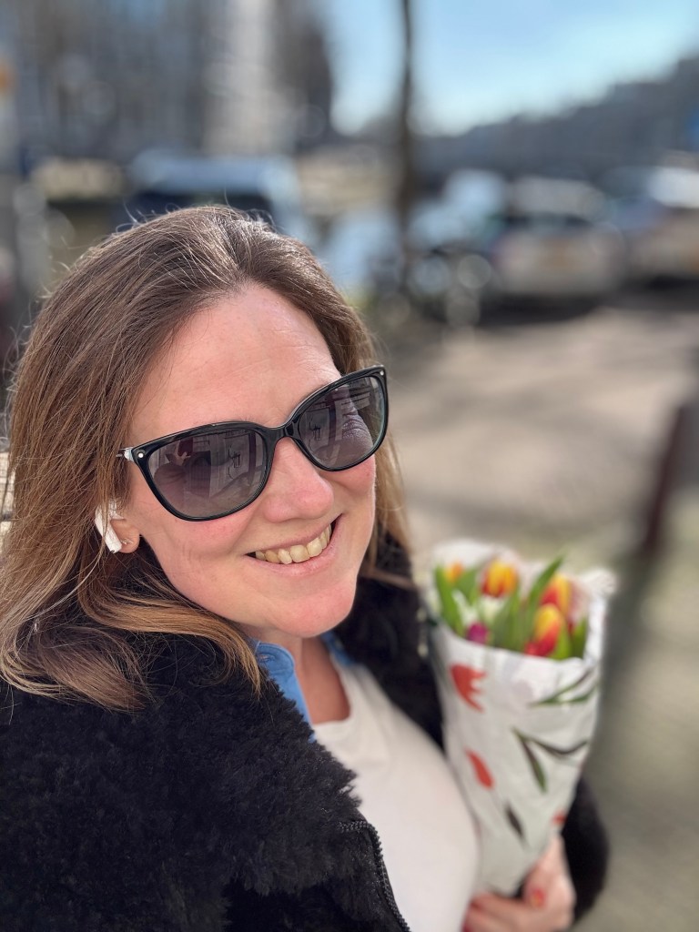 A smiling woman wearing sunglasses and wireless earbuds, holding a bouquet of tulips while standing outdoors with a city backdrop.