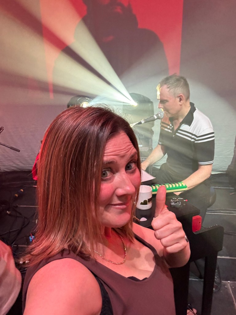 A woman giving a thumbs up while smiling in the foreground with a musician playing an instrument in the background under stage lighting.