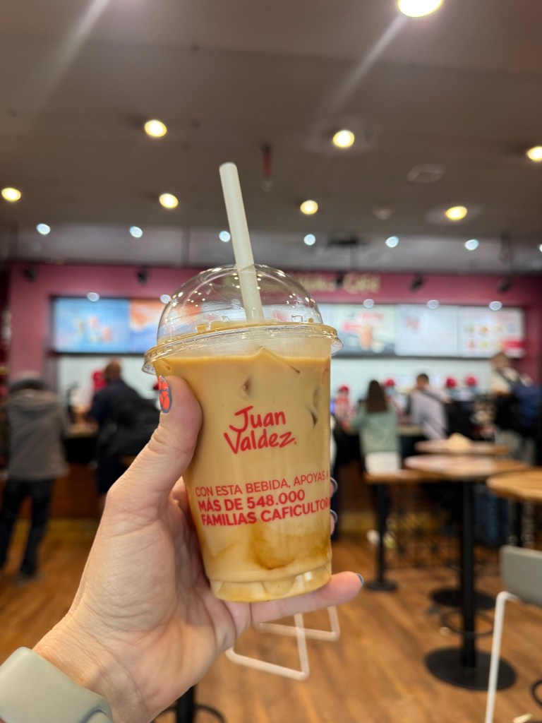 A person holding an iced coffee drink from Juan Valdez, featuring a clear cup with a dome lid and a straw, in a busy café setting.