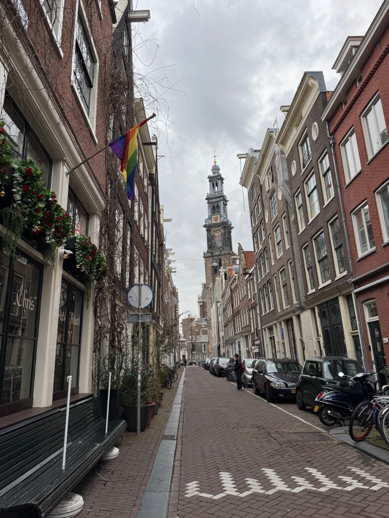 A narrow street in Amsterdam with a rainbow flag hanging, lined with traditional Dutch buildings, bicycles parked along the sides, and a church tower visible in the background under a cloudy sky.