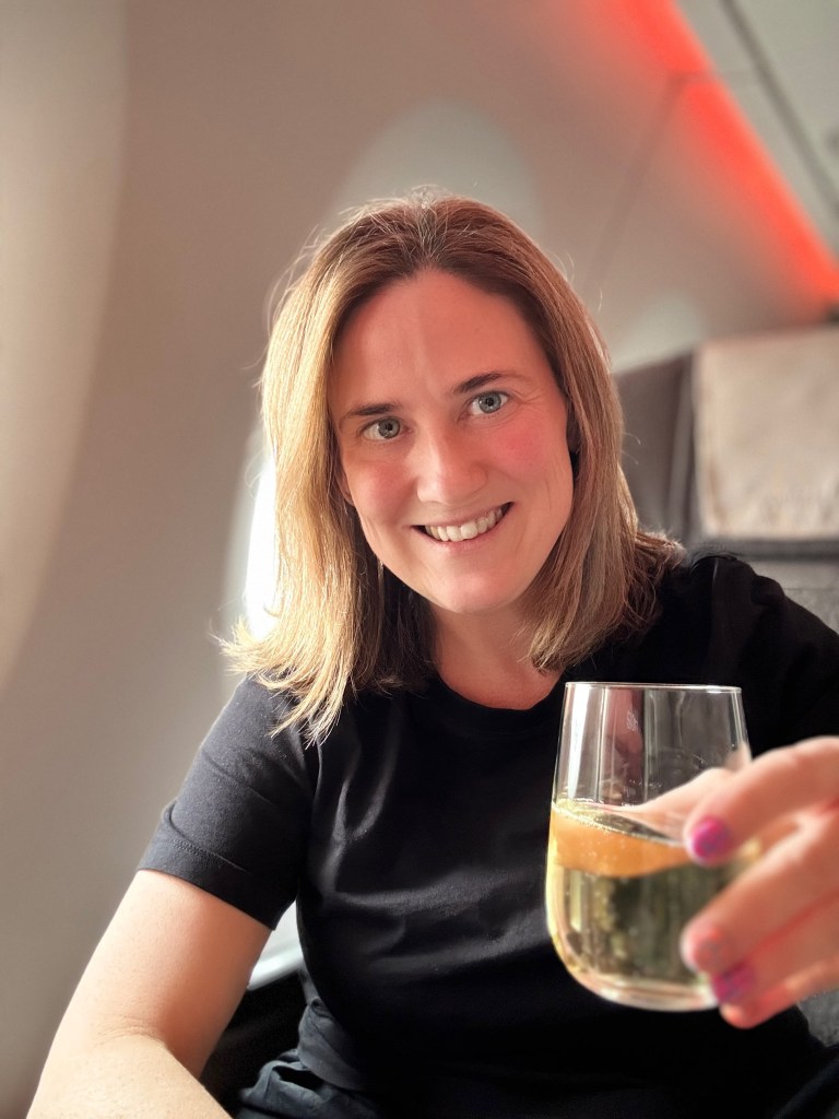 A person sitting on an airplane holding a glass of sparkling drink, smiling at the camera.