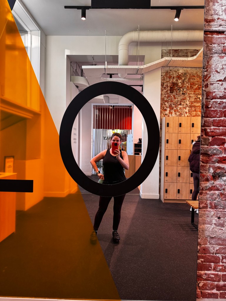 A person wearing a black tank top is taking a selfie in front of a circular mirror framed by a large black circle, with an orange geometric shape on the left. The background shows a gym interior with lockers and exercise equipment.