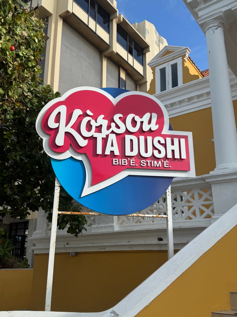 A colorful sign that reads 'Kòrsou Ta Dushi' with a heart design, located in front of a building surrounded by greenery.