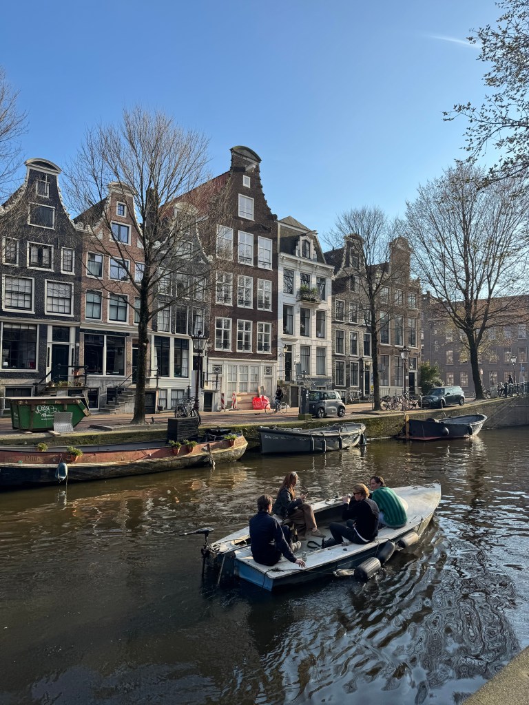 A group of people in a small boat on a canal, surrounded by historic buildings and trees under a clear blue sky.
