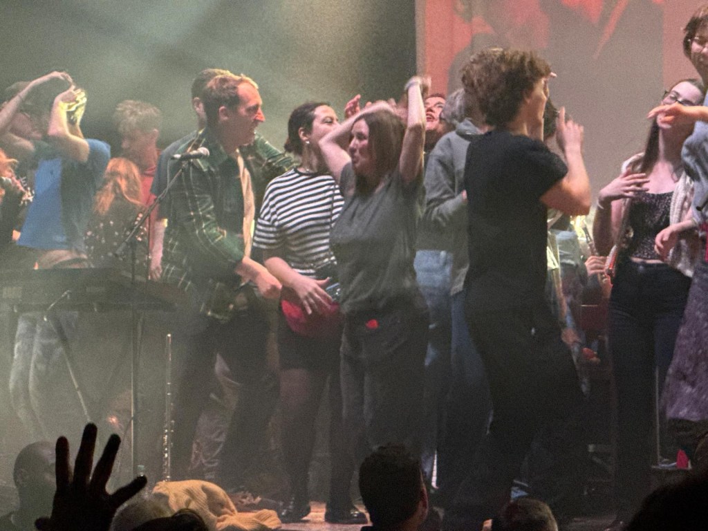 A crowd of people dancing enthusiastically on stage at a live music event, with a singer and musicians visible in the foreground, surrounded by colorful lights and a festive atmosphere.
