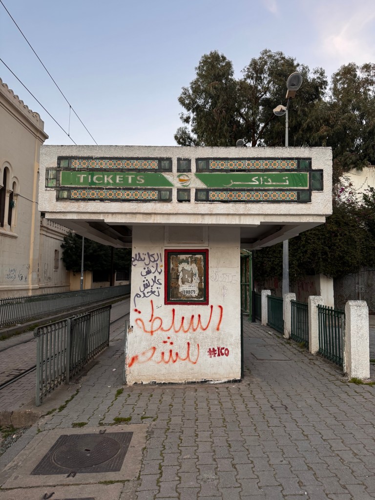 A weathered ticket booth with a green sign reading 'TICKETS' and Arabic text, surrounded by graffiti on the walls, located near tram tracks in an urban setting.