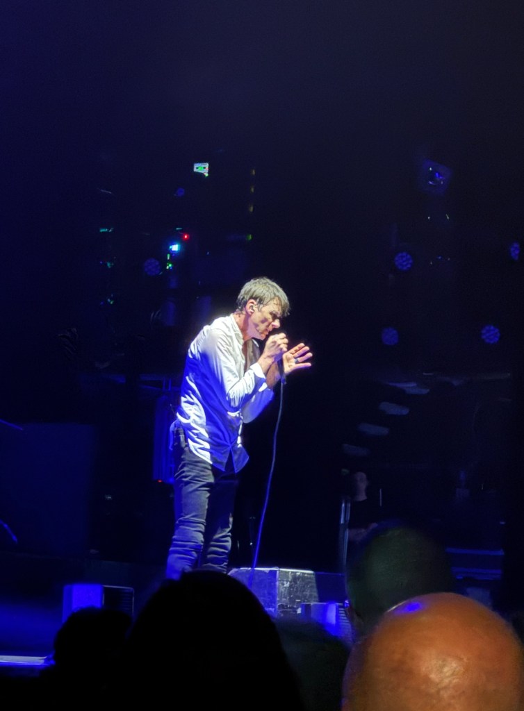 A singer performing on stage with a microphone, wearing a white shirt and black pants, surrounded by concert lights and an audience.