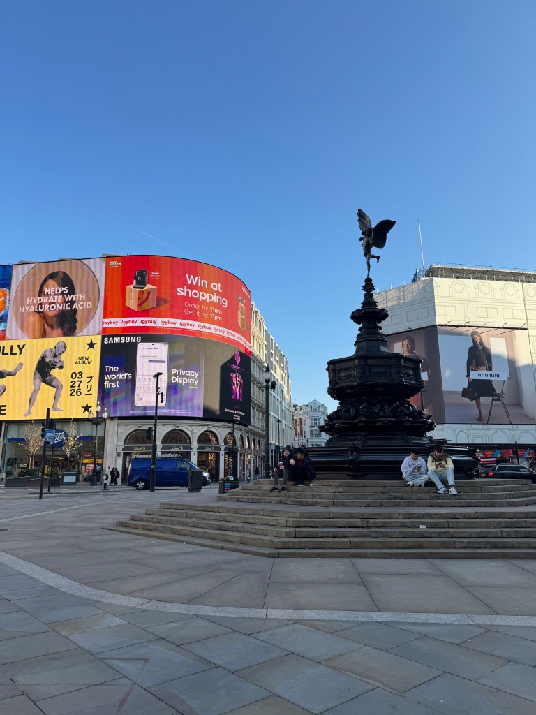 A view of Piccadilly Circus in London, featuring the iconic Eros statue, surrounded by various advertising billboards and people sitting on the steps.
