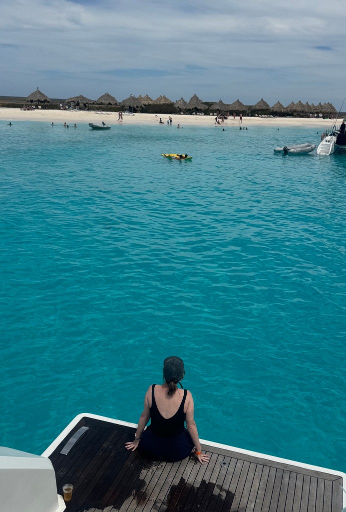 A person wearing a black swimsuit and cap sitting on a dock by crystal clear turquoise water, looking towards a beach with thatched umbrellas and people enjoying various activities.