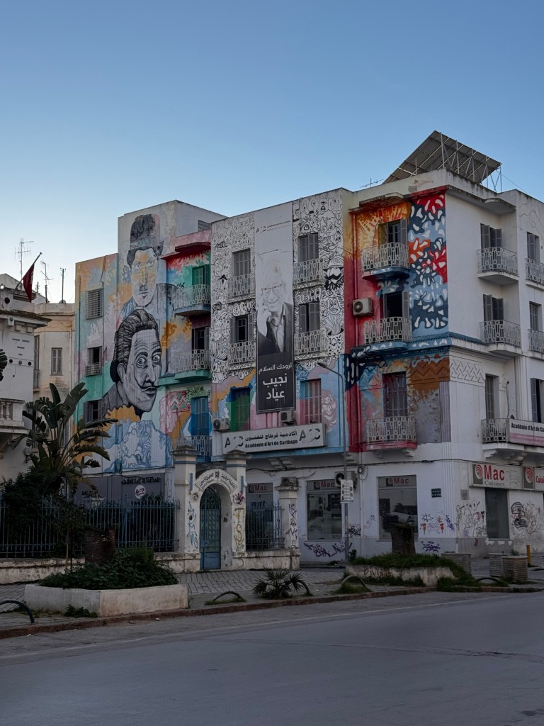 A colorful mural on the side of a building, featuring artistic portraits and abstract designs. The building has balconies and decorative elements, showcasing vibrant street art in an urban setting under a clear blue sky.