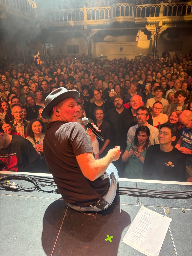 Stuart Murdoch from Belle and Sebastian sitting on stage with a microphone speaks to a large crowd of enthusiastic fans in a concert setting.