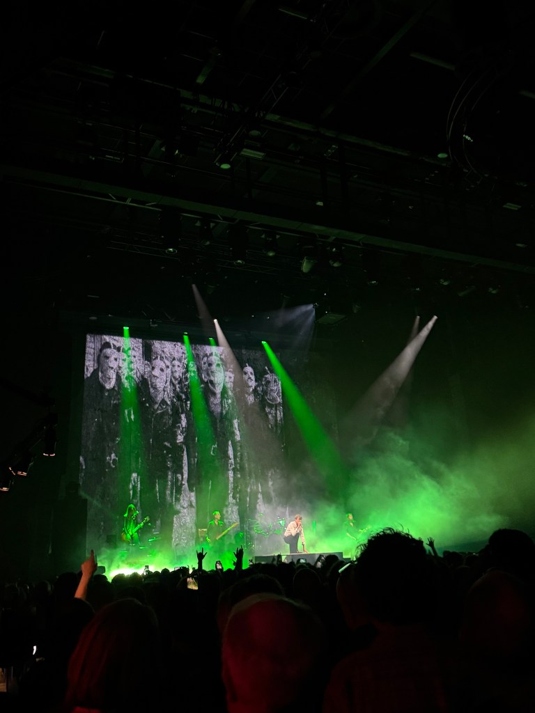A live concert scene with a band performing on stage, dramatic green lighting, and a large backdrop featuring black-and-white images of people. The audience is visible in the foreground, enjoying the performance.