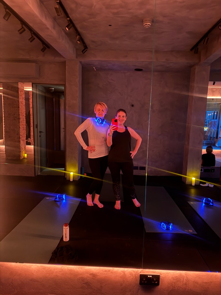 Two women posing in a fitness studio, standing in front of a mirror with yoga mats and workout equipment around them.