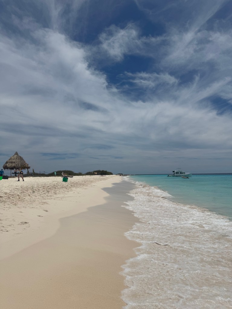 A scenic view of a tropical beach with soft white sand and gentle waves lapping at the shore, under a partly cloudy blue sky. A boat is visible in the clear turquoise water, and a thatched umbrella is placed on the beach.