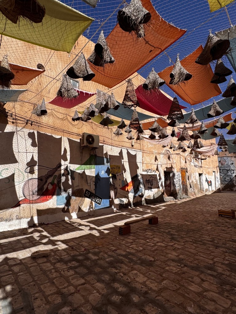 A colorful courtyard decorated with hanging woven baskets and vibrant fabric canopies, casting playful shadows on the cobblestone floor, featuring graffiti on the walls.
