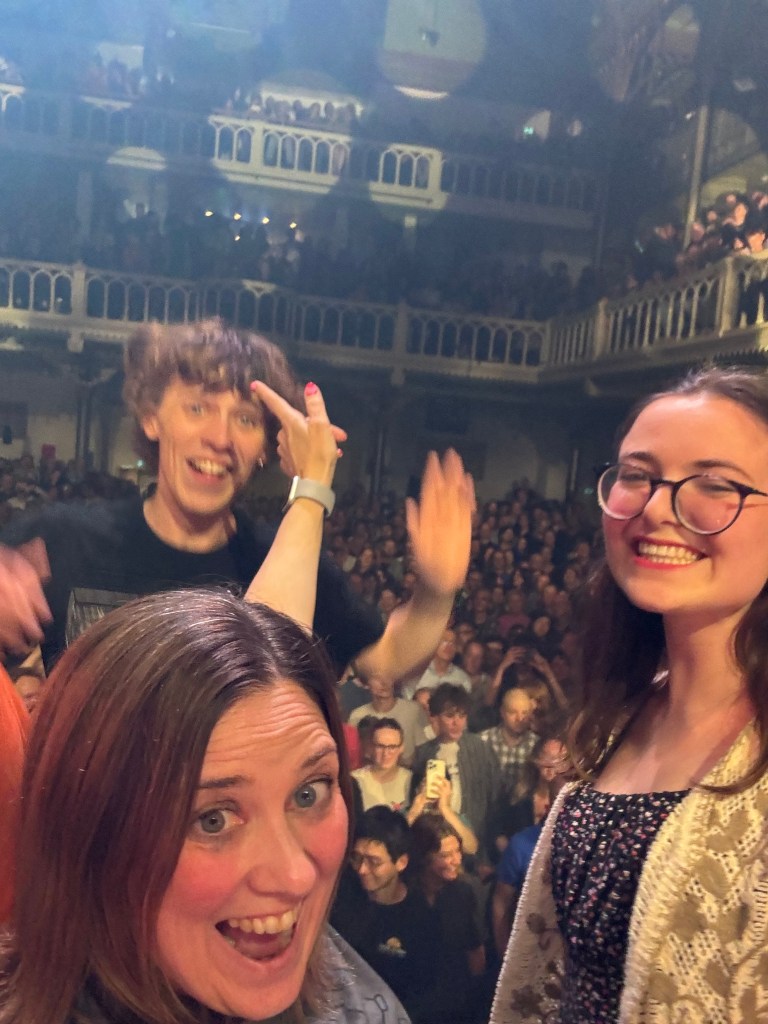 A group of three people smile and pose for a selfie at a concert, with a large crowd visible in the background.