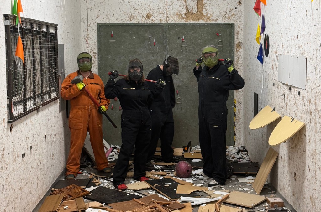 Four individuals wearing safety gear stand in a messy room filled with debris, holding various tools and equipment.