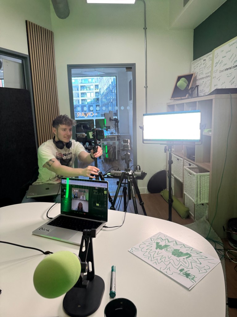 A young man sits in a studio setting, operating a camera on a tripod. In front of him is a laptop displaying a video call. A microphone and a green pen are placed on a round table, alongside a sheet of paper with a doodle design. The room features modern decor with large windows visible in the background.