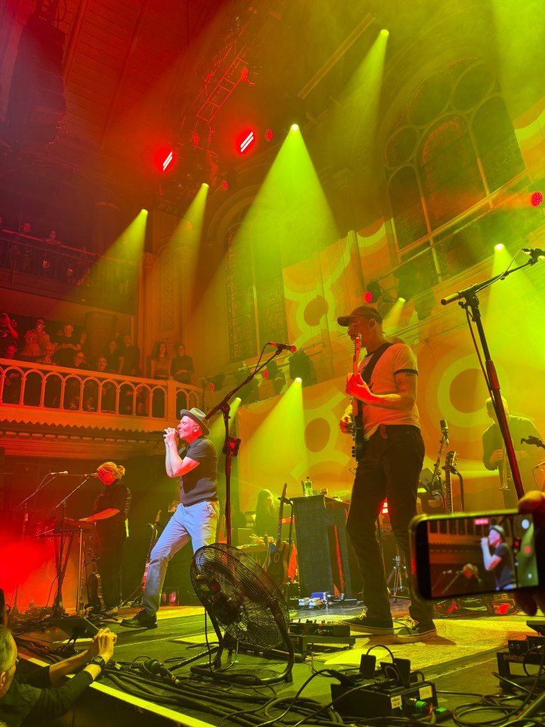 A live music performance with a band on stage, featuring vibrant lighting in red and green hues. The lead singer is singing passionately, while other band members play instruments. The audience is visible in the background, enjoying the show.
