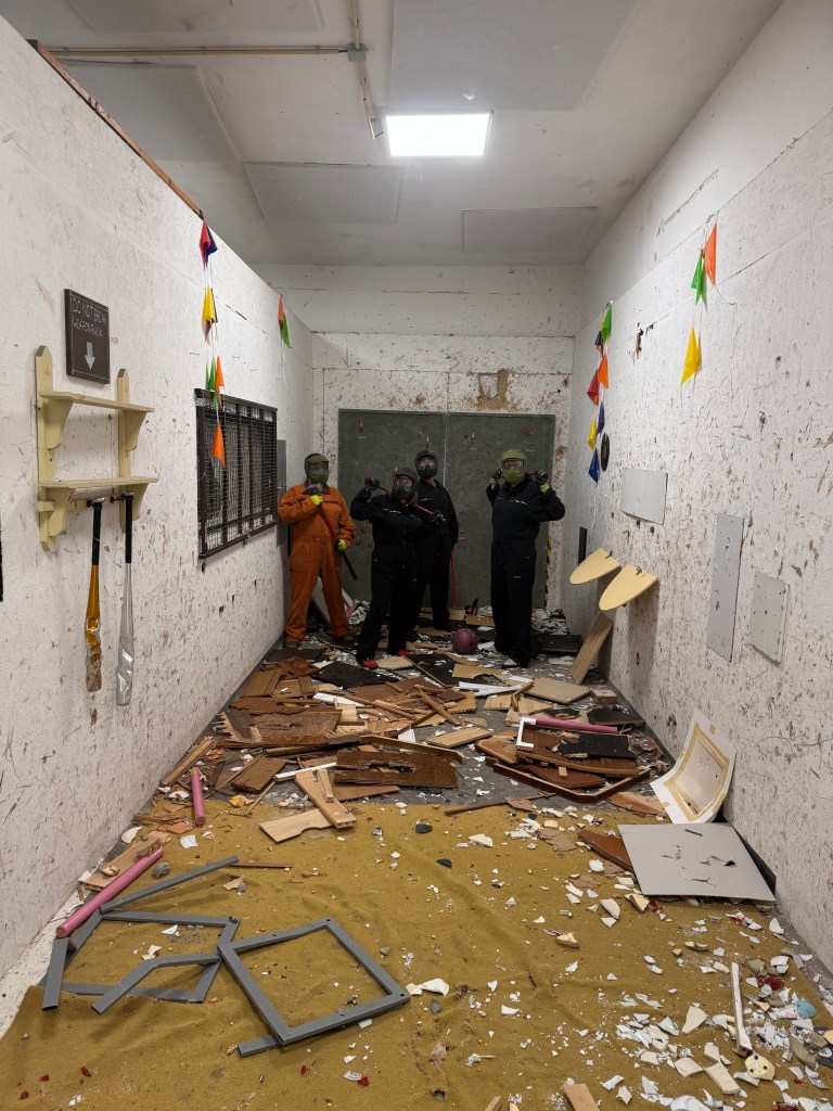 A chaotic hallway with debris scattered on the floor, featuring four individuals wearing masks and safety gear, posing for the camera. The walls are marked with damage, and colorful flags hang from the ceiling.