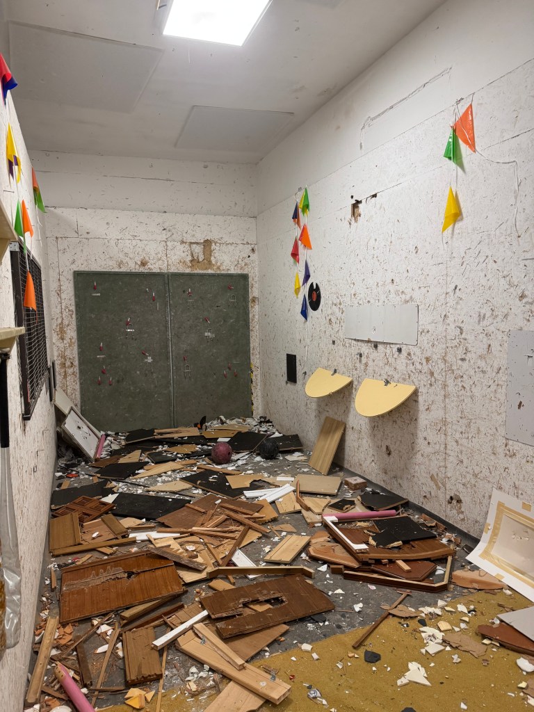 An untidy room with debris scattered across the floor, featuring broken wooden materials, a pink object, and colorful triangular decorations on the walls.