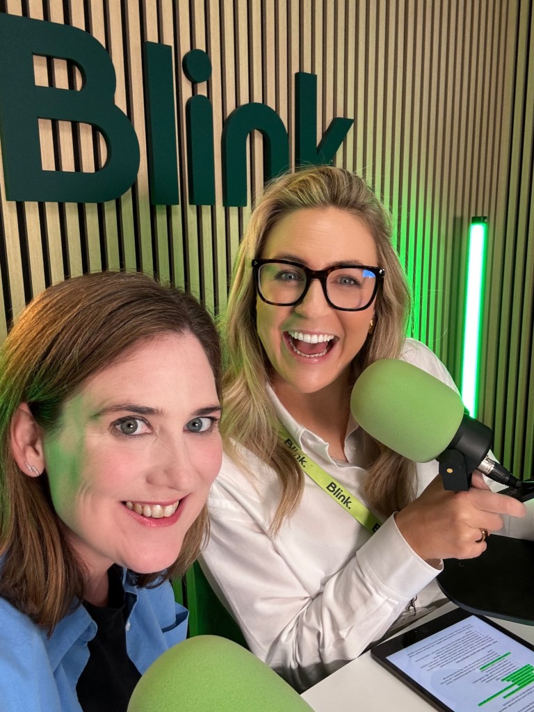 Sharon O'Dea and Georgie Scott are smiling at the camera in a studio setting, with a green 'Blink' sign in the background and green microphones in front of them.